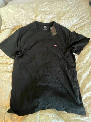 Svart t-shirt från Levi's - Klassisk svart t-shirt från Levi's med rund hals och liten röd logotyp på bröstet. Tillverkad i mjuk bomull och har en enkel, stilren design som passar till det mesta. Perfekt basplagg för garderoben.