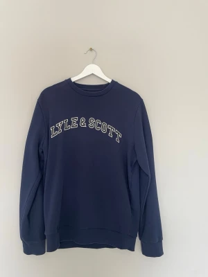 Mörkblå Hoodie från Lyle & Scott - Mörkblå sweatshirt från Lyle & Scott med vit logotyptext över bröstet. Klassisk rund halsringning och ribbade muddar vid ärmslut och nederkant. Perfekt för en avslappnad och stilren look. Pris kan diskuteras