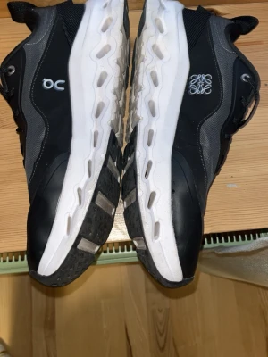 Svarta On Running sneakers - Snygga svarta sneakers från On Running med vita detaljer och tjock, vit sula. Skorna har meshpaneler för bra ventilation och logga på sidan. Perfekta för dig som gillar sportig och modern stil. Och de går o pruta