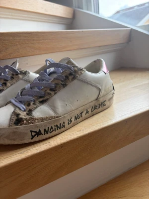 Vita sneakers med leoparddetaljer - Unika vita sneakers med leopardmönstrade detaljer och lila skosnören. Texten 'DANCING IS NOT A CRIME' syns på sidan av sulan och hälen har en rosa detalj. Skorna har en låg profil och coola glitterinslag vid tån. Perfekta för dig som vill sticka ut.