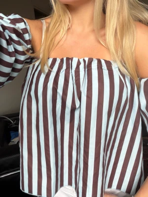 Randig offshoulder blus i blått och brunt - Säljer en trendig offshoulder blus med breda vertikala ränder i ljusblått och brunt. Blusen har puffiga ärmar och en lös, luftig passform som ger en cool och avslappnad vibe. Perfekt för dig som vill sticka ut med en färgglad och unik look.💙