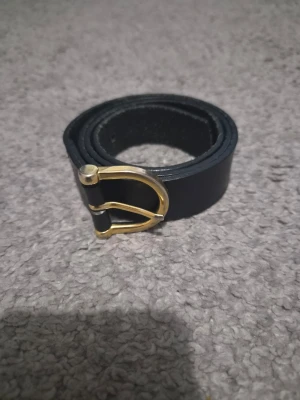 Svart skärp med guldfärgat spänne - Stilrent svart skärp i skinn med ett snyggt guldfärgat D-format spänne. Perfekt accessoar för att lyfta din outfit och passar till både jeans och byxor. Enkel design som funkar till många olika stilar.