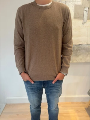 Beige stickad tröja med rund hals - En stilren beige stickad tröja med rund hals och långa ärmar. Tröjan har ribbade muddar vid ärmslut och nederkant. Perfekt att matcha med jeans för en avslappnad look. Materialet ser mjukt och bekvämt ut.