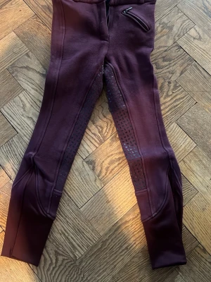Vinröda ridbyxor från CRW - Snygga vinröda ridbyxor från CRW med helskoning i silikon för extra grepp. Byxorna har en dragkedjeficka fram och stretchigt material som ger bra rörelsefrihet. Perfekta för ridning och träning. Lite krympande i tvätten så lite mindre än 130