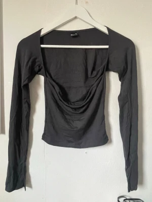 Grå långärmad topp Gina Tricot - Snygg svart långärmad topp från Gina Tricot med draperad front och fyrkantig urringning. Toppen har en tight passform och är tillverkad i mjukt material som sitter skönt på kroppen. Perfekt att styla med jeans eller kjol.