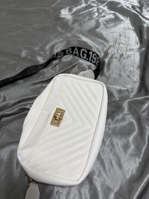 Vit axelväska med textad rem - Snygg vit axelväska i quiltad design med guldfärgat spänne framtill. Väskan har en bred, svart axelrem med vit text där det står 'BAG.1980' och andra ord. Perfekt för dig som vill ha en trendig och praktisk accessoar. Materialet ser ut att vara konstläder.