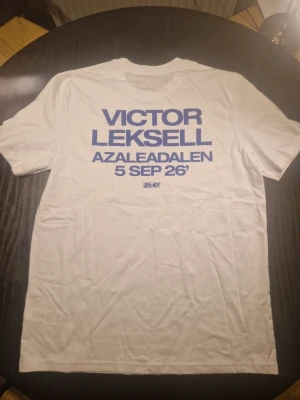 Vit t-shirt Victor Leksell Azaleadalen - Säljer en vit t-shirt med stor blå text på ryggen: 'Victor Leksell Azaleadalen 5 Sep 26'. På framsidan finns ett stort blått blommotiv med rosa kant. T-shirten har korta ärmar och är i mjuk bomull, perfekt för dig som gillar konsertmerch och snygga prints.