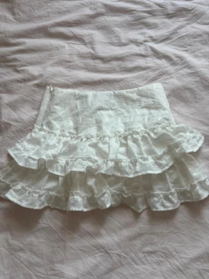 Vit volangkjol H&M - Söt vit minikjol med dubbla volanger och broderade detaljer. Kjolen är gjord i ett lätt bomullstyg som ger en romantisk och luftig känsla. Perfekt för dig som vill ha en feminin och trendig stil.