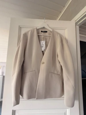 Beige kavaj från Gina Tricot - Stilren beige kavaj från Gina Tricot med en knapp framtill och två fickor. Kavajen har en rak passform och är tillverkad i ett mjukt, vävt material. Perfekt för en clean och modern look. Lappen kvar och aldrig används