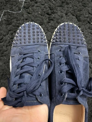 Mörkblå sneakers med nitar - Snygga mörkblå sneakers med matta nitar över tån och klassisk röd sula. Skorna har snörning och är tillverkade i mocka för en lyxig känsla. Perfekta för dig som vill sticka ut med en edgy stil.