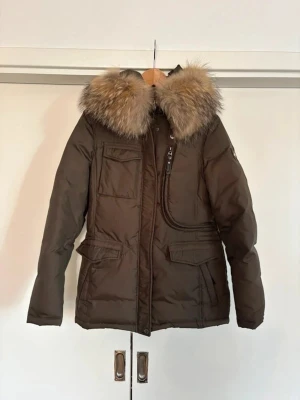 Brun dunjacka med pälskrage - Snygg brun dunjacka med stor fluffig pälskrage och huva. Jackan har flera fickor framtill, dragkedja och tryckknappar. Perfekt för kalla vinterdagar och har en klassisk, lite längre modell.