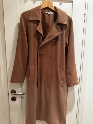 Klassisk brun trenchcoat med bälte - Stilren brun trenchcoat med dubbelknäppning, bred krage och knytskärp i midjan. Jackan har raka ärmar och två fickor framtill. Perfekt för dig som vill ha en tidlös och snygg look. Materialet ser ut att vara bomull eller en bomullsblandning.