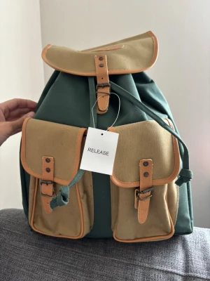 Release Grön och beige canvasryggsäck - Snygg Realise ryggsäck i grön och beige canvas med detaljer i brunt konstläder. Två stora ytterfickor framtill med spännen och justerbara axelband. Stängs med dragsko och lock med spänne. Insidan är vattenavvisande. Perfekt för skola eller utflykter. Mått 34x40x18 cm 