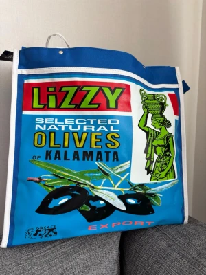 Blå shoppingväska med olivtryck - Färgglad shoppingväska från Daniel Ray med mexikanskt inspirerat tryck.  Väskan har vita handtag och kantband samt rymlig rektangulär form. Perfekt för att bära med sig allt man behöver till skolan, stranden eller stan. Mått 39x36x11 cm 