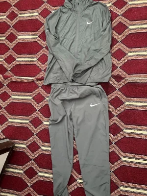 Grå nike Miller set - Säljer ett grått träningsset från Nike med matchande jacka och byxor. Setet har en sportig look med vita Nike-loggor på både jackan och byxorna. Materialet är lätt och passar perfekt för träning eller outfit. Pris kan diskuteras 