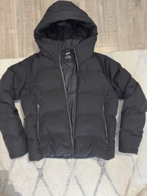 Svart pufferjacka från Uniqlo - Säljer en svart pufferjacka från Uniqlo i storlek M. Jackan har huva, två dragkedjeförsedda fickor och är tillverkad i ett vattenavvisande och lätt material. Perfekt för kalla vinterdagar och har en stilren, boxig passform.