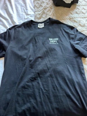 Svart t-shirt från Gallery Dept. - Säljer en svart t-shirt från Gallery Dept. med tryck i vitt på både bröstet och ryggen. Klassisk rund halsringning och korta ärmar. Perfekt för dig som gillar streetwear och vill sticka ut med en stilren look.