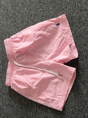 Ljusrosa shorts med snörning - Snygga ljusrosa shorts med elastisk midja och vit snörning. Shortsen har en liten blå logga broderad framtill och en bakficka med knapp. Perfekta för sommardagar och har en avslappnad passform.