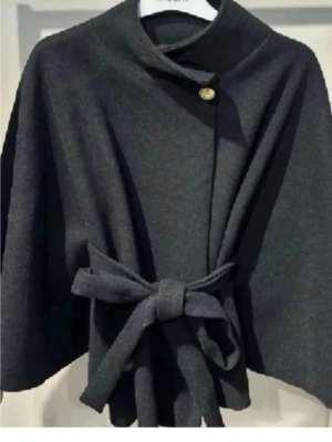Säljer denna trendiga kappan  - Stilren svart kappa med hög krage, dekorativ guldknapp och knytband i midjan. Kappan har vida ärmar och en elegant, enkel design som passar perfekt till höst och vår. Storlek one size