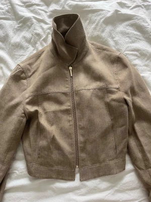 Beige cropped jeansjacka - Snygg beige jeansjacka i croppad modell med dragkedja framtill. Jackan har två olika kragar – en hög ståkrage och en klassisk skjortkrage, vilket ger en unik look. 