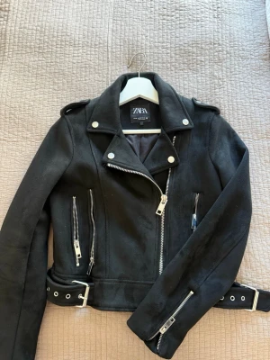 Svart jacka från ZARA - Snygg svart jacka från ZARA med silvriga dragkedjor, axelklaffar och bälte i midjan. Jackan har flera fickor med dragkedja och klassisk krage. Perfekt för en cool och trendig look. Materialet är som sammet. Använd ett fåtal gånger! 