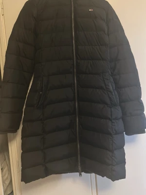 Svart lång dunjacka med fuskpäls - Snygg svart lång dunjacka med horisontella sömmar och dragkedja framtill. Jackan har två fickor med dragkedja och en huva med grå fuskpäls. Perfekt för kalla vinterdagar och ger en stilren look. Endast använt en vinter säsong. 