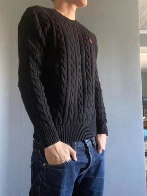 Ralph Lauren kabelstickad - Snygg svart kabelstickad tröja med rund hals ribbade muddar vid armslut och vid nedrekant från Ralph Lauren. Tröjan har ett broderat märke på bröstet och den är perfekt vid lager på lager. Passar både till jeans men också om man vill klä upp sig lite så passar de perfekt med chinos tex. Det finns ingen lapp kvar men den är knappt använd och jag har kvar plast påsen man får den i när man köper den ny. Man kan pruta ner priset vid snabb affär men jag vill helst inte få några skambud tack!