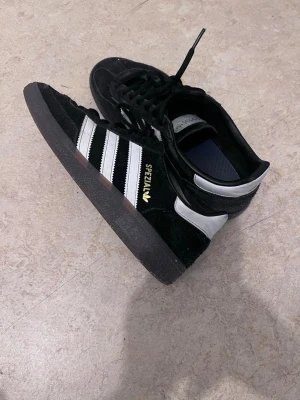 Svarta Adidas Spezial sneakers - Klassiska Adidas Spezial sneakers i svart mocka med vita detaljer och de ikoniska tre ränderna på sidan. Guldfärgad 'SPEZIAL'-text på sidan och svart sula. Skorna har snörning och låg profil. Perfekta för dig som gillar retrostil och streetwear.