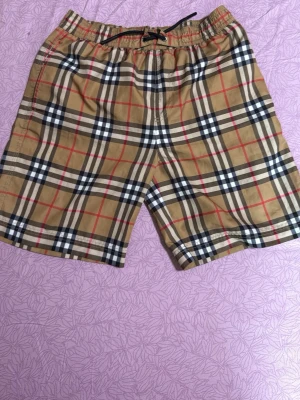 Rutiga beige shorts med snörning - Snygga shorts i klassiskt beige med rutig design i svart, vitt och rött. De har elastisk midja med snörning och är perfekta för sommaren. Materialet känns lätt och bekvämt, och shortsen har en avslappnad passform.