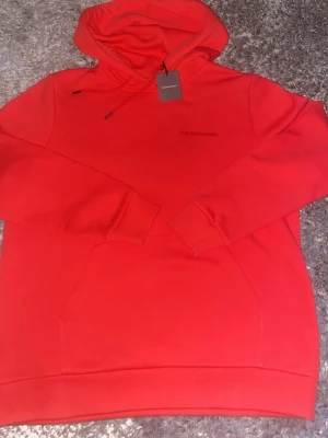 Röd hoodie från Peak Performance - Snygg röd hoodie från Peak Performance med broderad logga på bröstet. Klassisk känguruficka framtill och justerbar huva med dragsko. Tillverkad i mjukt material som känns skönt mot huden. Perfekt för en avslappnad och trendig stil.
