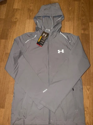 Grå vindjacka från Under Armour Passar storlek S -  De står Storlek M i tröjan men passar igentligen SSnygg grå vindjacka från Under Armour med huva och dragkedja framtill. Jackan har vita detaljer, logga på bröstet och praktiska sidofickor. Tillverkad i lätt och slitstarkt material, perfekt för blåsiga dagar. Sportig look med reflekterande detaljer.
