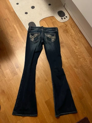 Mörkblå bootcut jeans med brodyr - Säljer ett par mörkblå bootcut jeans med snygg broderad design på bakfickorna. Jeansen har kontrastsömmar och klassisk femficksmodell. Perfekta för dig som gillar en lite retro vibe och vill sticka ut med detaljer. Materialet är denim.