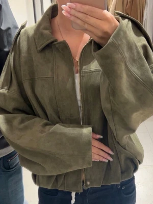 Mockajacka Zara - Säljer en olivgrön oversized jacka i mocka-look med dragkedja framtill och bred krage. Jackan har långa ärmar och en avslappnad passform som ger en cool och trendig vibe. Perfekt att slänga över en t-shirt och jeans.