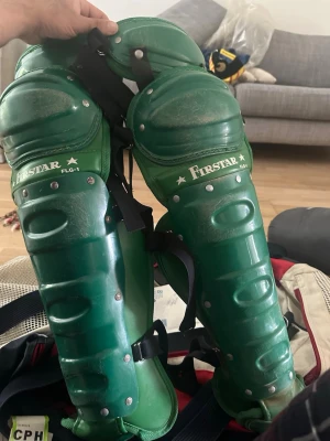 Gröna benskydd från Firstar - Säljer ett par gröna benskydd från Firstar, modell FLG-1. Skydden har justerbara svarta remmar och är tillverkade i hårdplast med metallnitar för extra hållbarhet. Perfekta för sporter där extra benskydd krävs.