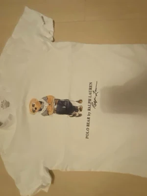 Vit Polo Bear t-shirt från Ralph Lauren - Vit t-shirt från Ralph Lauren med ikoniskt Polo Bear-tryck på bröstet. Björnen har randig tröja och solglasögon, vilket ger en cool vibe. Klassisk passform och rund hals. Perfekt för dig som gillar streetstyle och vill ha något tidlöst.