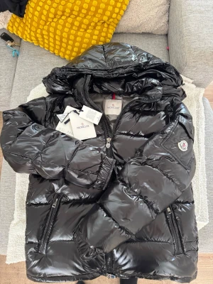  Moncler Maya  - Säljer en svart glansig dunjacka från Moncler med huva och dragkedja framtill. Jackan har två fickor med dragkedja och Moncler-logga på ärmen. Perfekt för kalla vinterdagar och riktigt snygg streetstil. Stl L sälja för den inte passar mig. Det står den 1 som stl men det stämmer ej. Tags medföljer ej använd. 