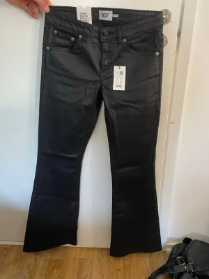 Svarta bootcut jeans från 157 - Snygga svarta bootcut jeans från Lager 157 med låg midja och fem klassiska fickor. Jeansen har en lätt glansig finish som ger en cool look och är perfekta för att lyfta vilken outfit som helst. 