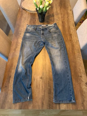 Blå raka jeans med slitningar - Säljer ett par klassiska blå jeans med raka ben och lätt slitna detaljer. Jeansen har fem fickor, normal midja och dragkedja med knapp. Materialet är denim i bomull och färgen är tvättad blå med ljusare partier för en avslappnad look.