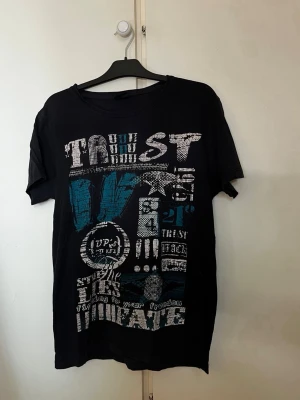 Svart/blå t-shirt med grafiskt tryck - Cool svart t-shirt med stort grafiskt tryck i vitt och blått framtill. T-shirten har rund hals och korta ärmar. Perfekt för dig som gillar streetwear och vill sticka ut lite extra.