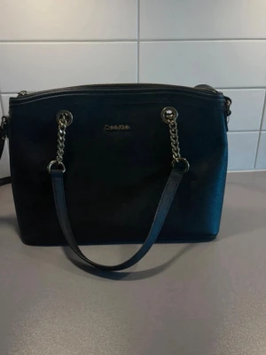 Svart handväska från Calvin Klein - Välj gärna instabox, kan skicka mycket snabbare då :). Mycket fint skick på väskan, använd 1 gång. Snygg svart handväska från Calvin Klein i stilren design. Väskan har guldfärgade kedjedetaljer och logga framtill. 