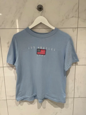 Ljusblå t-shirt med USA-flagga - Ljusblå t-shirt med broderad amerikansk flagga och texten 'Los Angeles 1992' i strl S/36. Klassisk rund halsringning och korta ärmar. Perfekt för en avslappnad och cool stil🩵