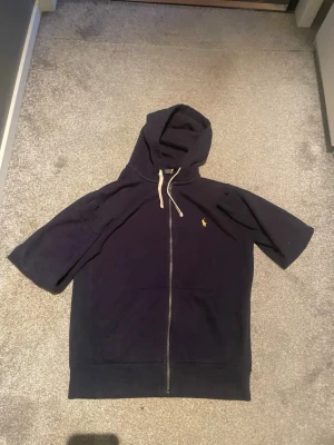 Mörkblå hoodie med dragkedja - Säljer en mörkblå hoodie med dragkedja. Hoodien har en huva med vita snören och en liten gul logga på bröstet. Praktiska fickor framtill och avslappnad passform.