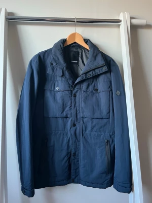 J Lindeberg fieldjacket mörkblå - Stilren mörkblå fieldjacket med krage och flera praktiska fickor framtill. Jackan har både knappar och dragkedja samt lång ärm. Perfekt för blåsiga dagar och enkel att matcha med olika outfits.