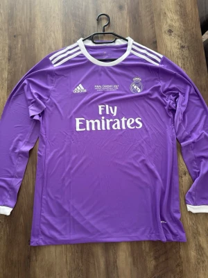 Lila Real Madrid långärmad matchtröja - Säljer en lila långärmad Real Madrid matchtröja från Adidas med vita detaljer och tryck. Tröjan har tryck för Champions League-finalen 2017 i Cardiff, klubbmärke på bröstet och Fly Emirates som sponsor. Perfekt för dig som älskar fotboll och Real Madrid.