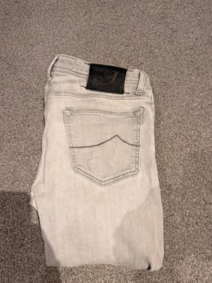 Ljusgrå jeans från Jacob Cohen - Snygga ljusgrå jeans från Jacob Cohen med klassisk femficksdesign och broderad J-detalj på myntfickan. Jeansen har raka ben och är tillverkade i mjukt denimtyg med en svart patch bak i midjan. Dem är väl använda med tecken på användning (se bilder). Släpper dem därav väldigt billigt, nypris runt 4000kr (väger 88 och är 190 och dem är små) 