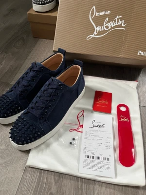 Mörkblå Christian Louboutin sneakers - Snygga mörkblå sneakers från Christian Louboutin med klassiska röda sulor och coola nitar framtill. Skorna är låga och har ovandel i mocka med vita gummisulor. Perfekta för dig som vill sticka ut med en lyxig och edgy stil.