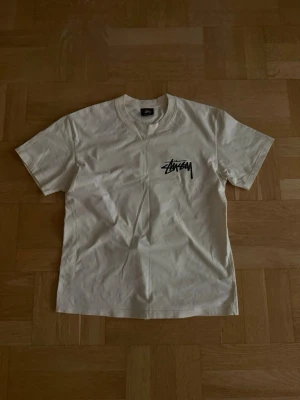 Vit Stüssy t-shirt med blå tärningar - Vit t-shirt från Stüssy med svart logga på bröstet och stor logga samt blå tärningar på ryggen. Klassisk passform med rund hals och korta ärmar. Perfekt för dig som gillar streetwear och vill sticka ut med en cool design.