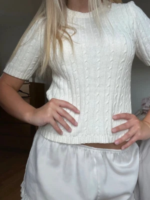 Vit kabelstickad tröja Gina Tricot - Kabelstickad vit tröja från Gina Tricot med korta ärmar och rund halsringning. Tröjan har en klassisk, figurnära passform och ribbade muddar vid ärmslut och nederkant. Perfekt till jeans eller kjol för en clean look.