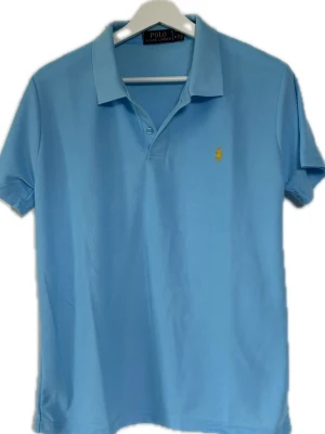 Ljusblå pikétröja från Polo Ralph Lauren - Klassisk ljusblå pikétröja från Polo Ralph Lauren med korta ärmar och krage. Tröjan har två knappar vid halsen och den ikoniska gula logotypen broderad på bröstet. Perfekt till jeans eller shorts för en clean look. ‼️ALDRIG ANVÄND‼️