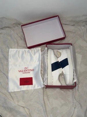 Valentino open - Snygga vita sneakers från Valentino Garavani med en bred mörkblå detalj över snörningen. Skorna är tillverkade i läder och har klassisk låg profil med vita skosnören och diskret logotyp på plösen. Perfekt för dig som gillar stilrena och exklusiva sneakers.